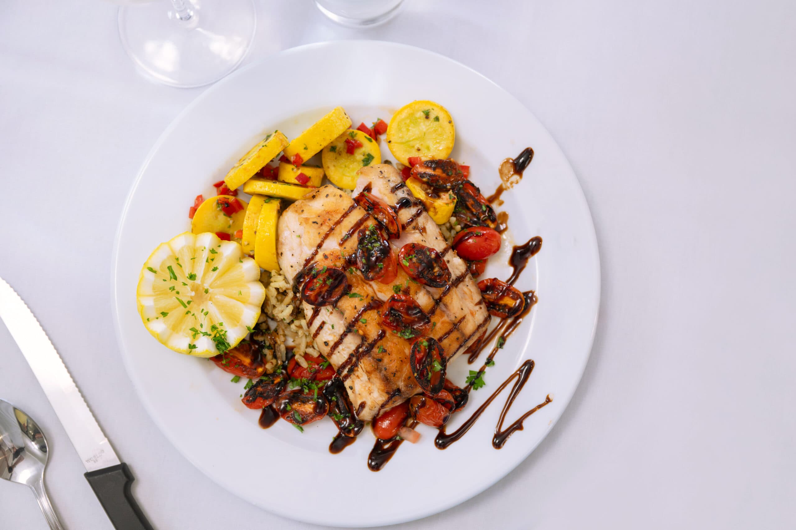 Overhead view of a white plate holding a grilled chicken breast topped with cherry tomatoes, served alongside yellow squash and a balsamic glaze drizzle.