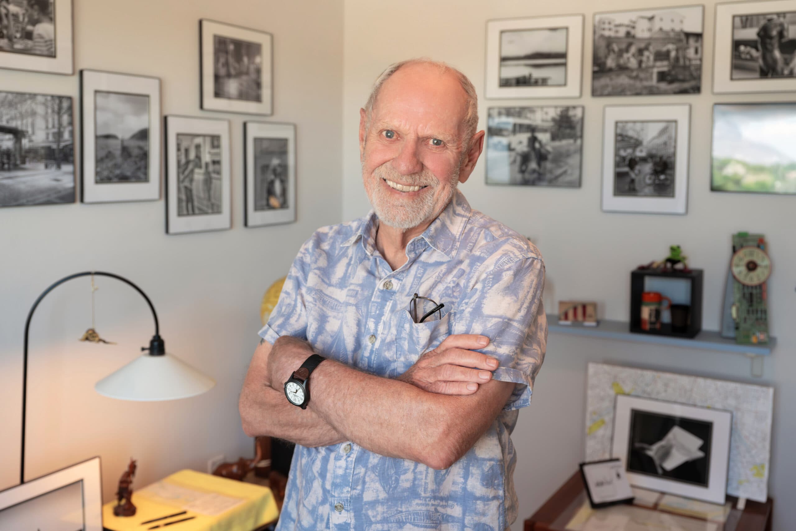 An elderly man standing in a living room filled with framed photos, arms crossed and smiling warmly at the camera.