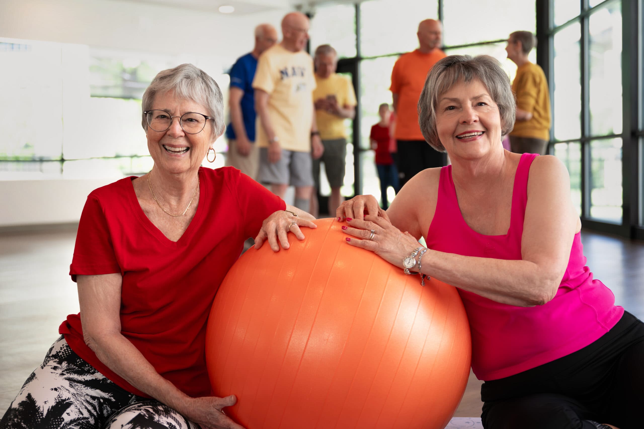 An elderly woman and a female trainer standing side by side in a gym, both flexing their arms and celebrating with big smiles.