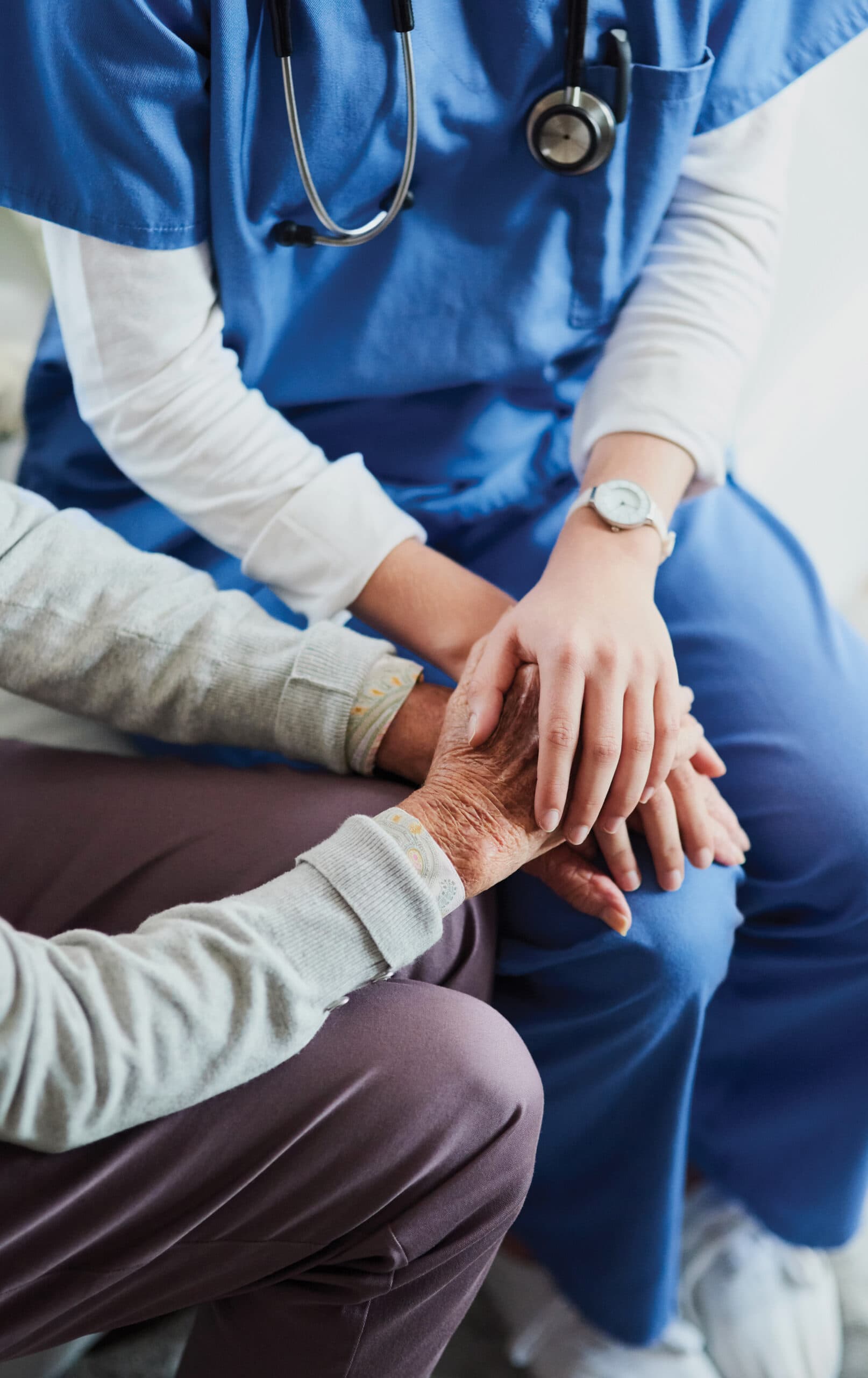 Close-up of a nurse in blue scrubs placing her hand reassuringly over an elderly patient’s hand during memory care services.