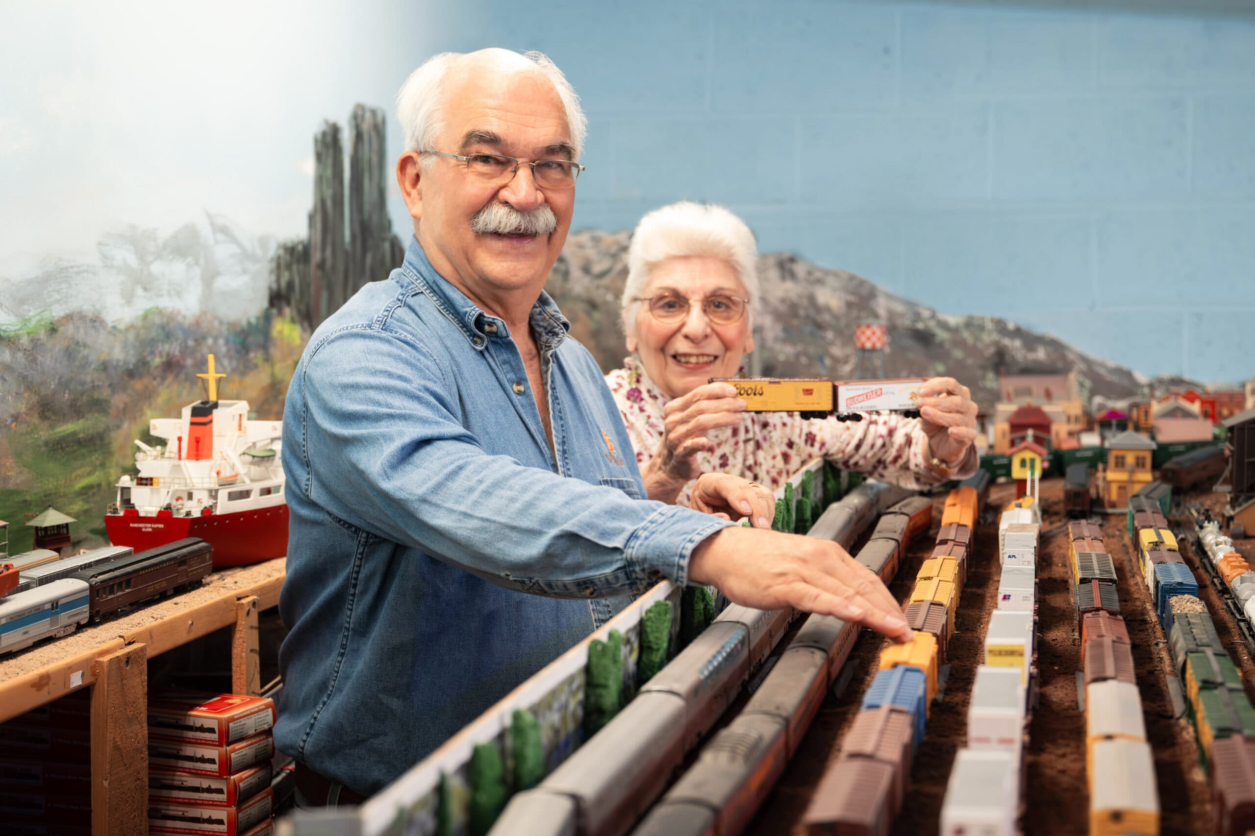 An elderly man and woman arranging model train cars on a detailed indoor railroad layout at a hobby table.