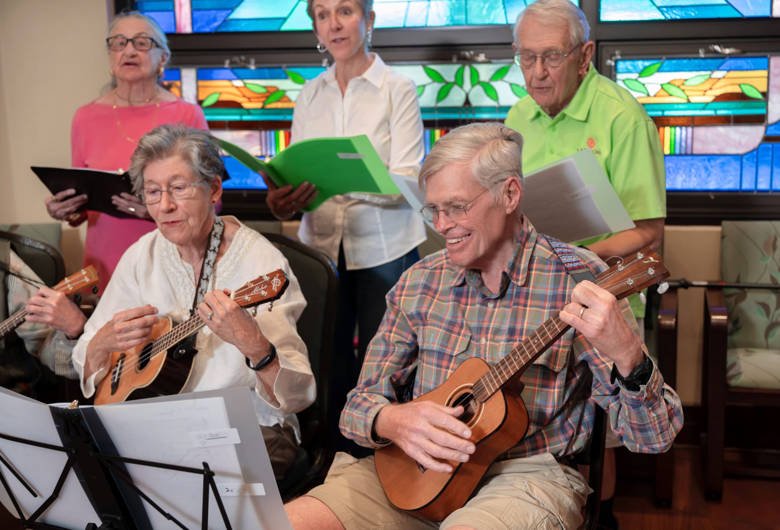 A small ensemble of seniors playing ukuleles in front of a standing choir singing in a room with stained glass windows.
