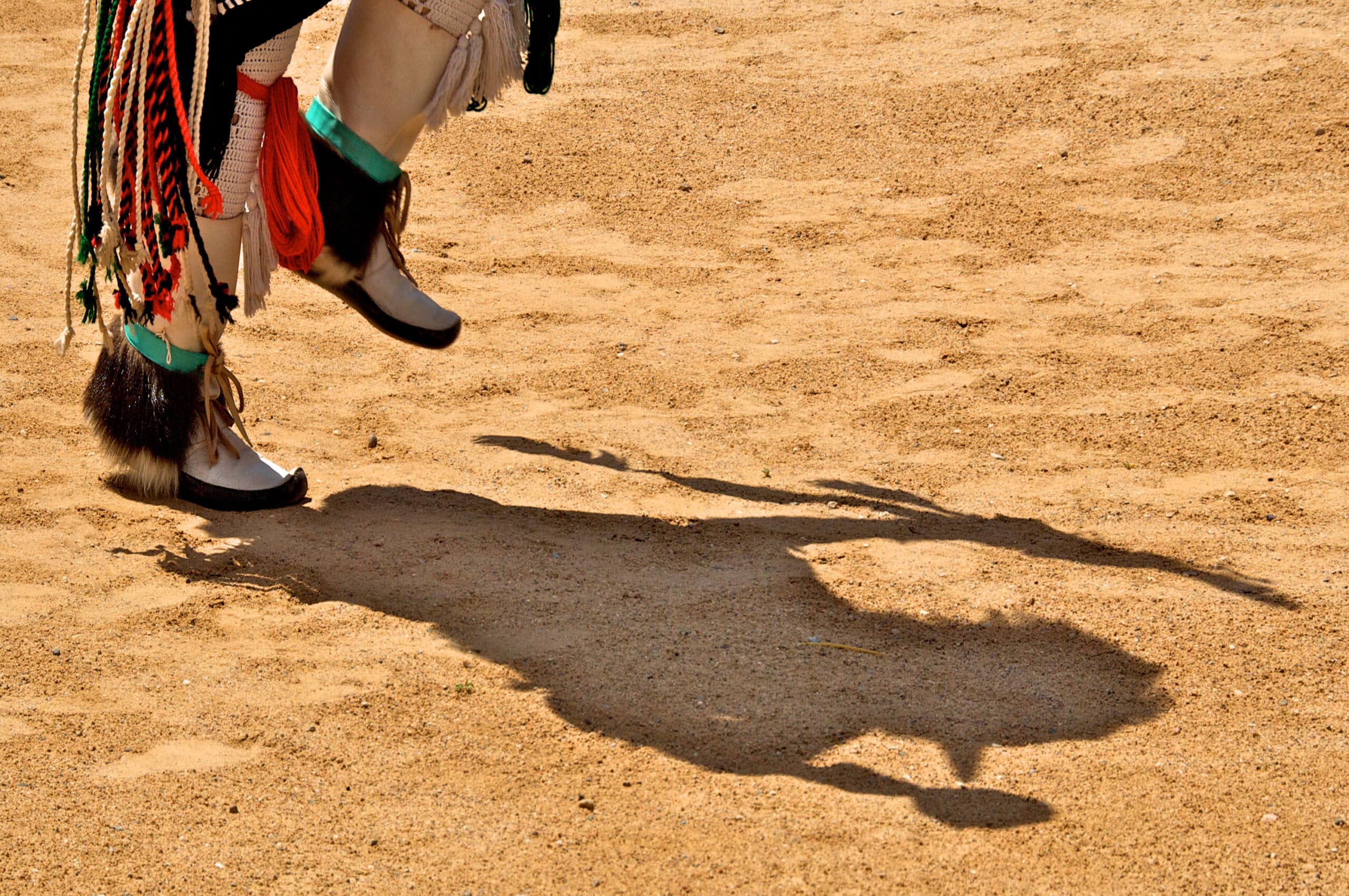 Close-up of Native American dance regalia—feathered fringe and beadwork on moccasined feet—casting a shadow as the dancer’s foot lifts off the sandy ground.