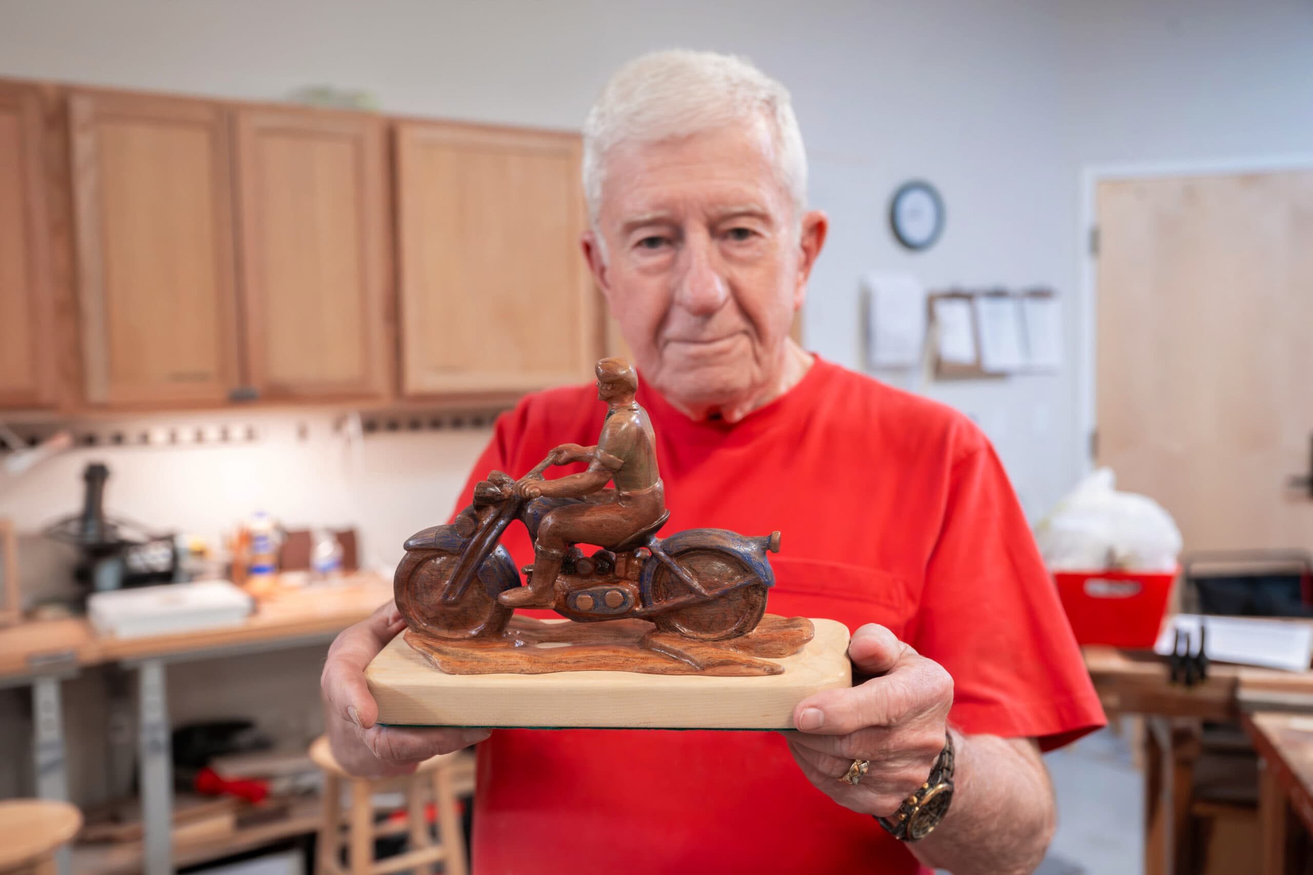 An older man in a red shirt holding up a carved wooden sculpture of a motorcyclist on a bike, displaying it proudly in a woodshop.