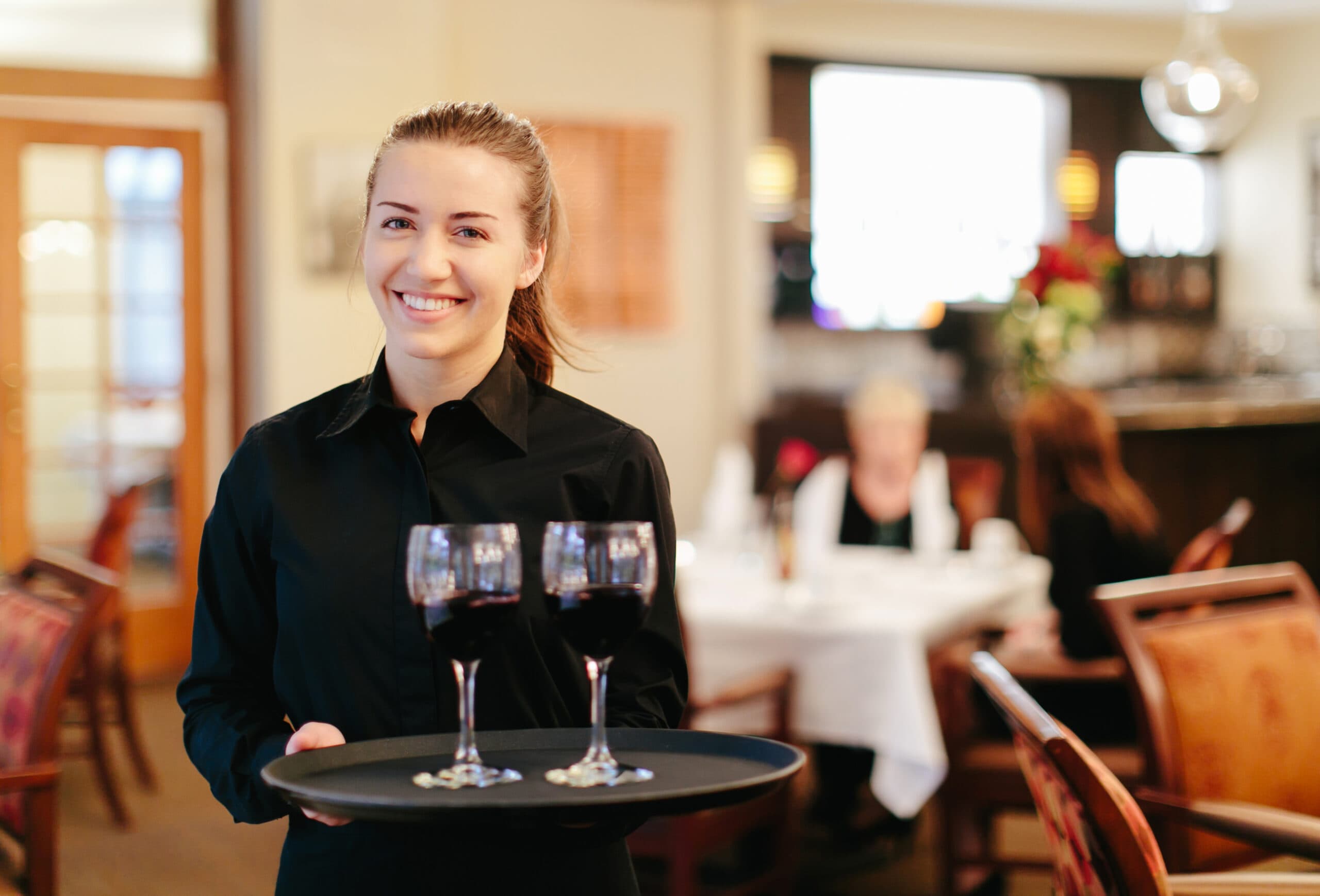 A smiling server in a black shirt carrying a round tray with three glasses of red wine in a warmly lit dining room.