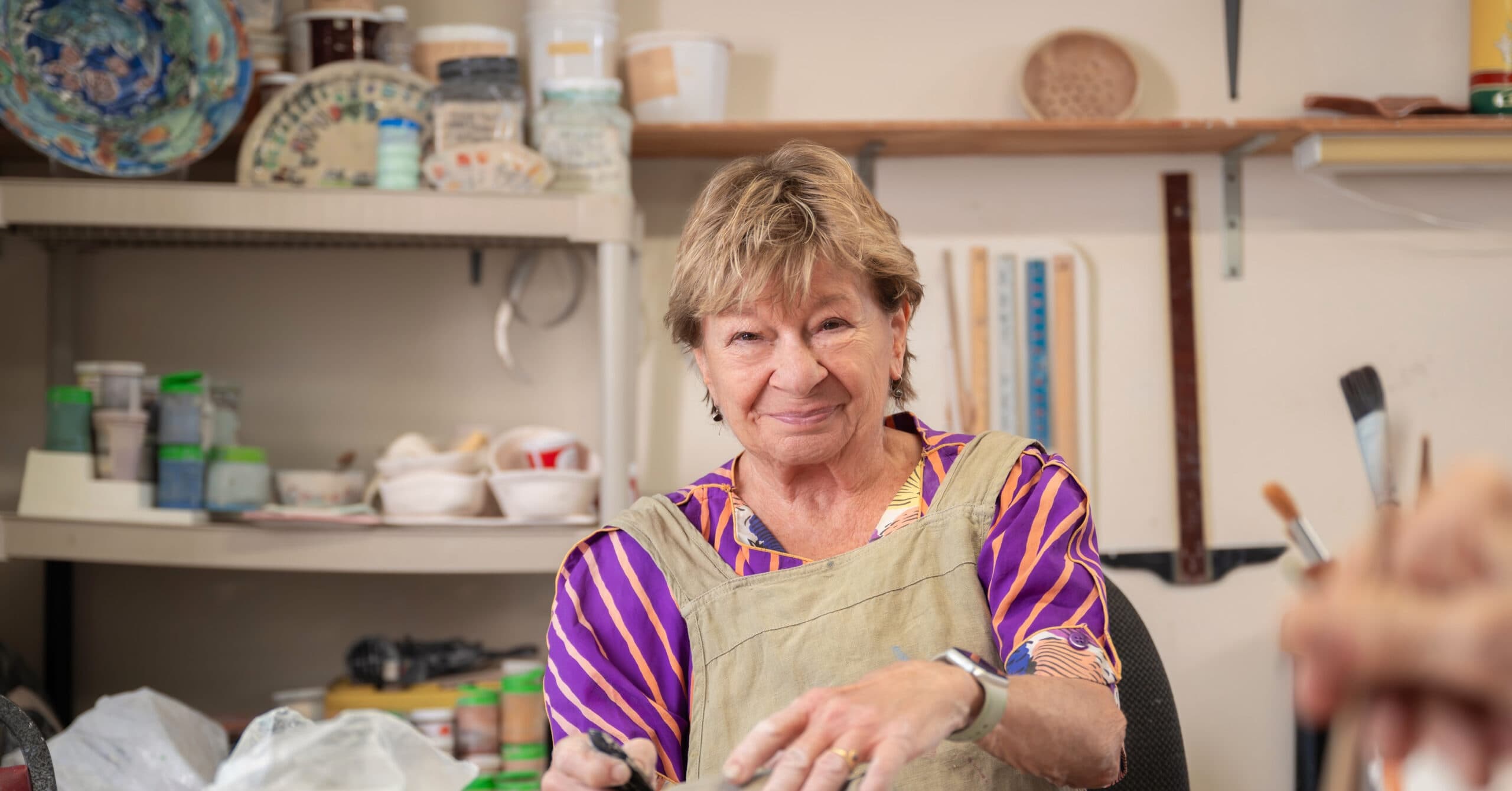 An elderly woman sitting at a pottery studio table, shaping a lump of clay with a tool, surrounded by shelves of glazes and ceramics.